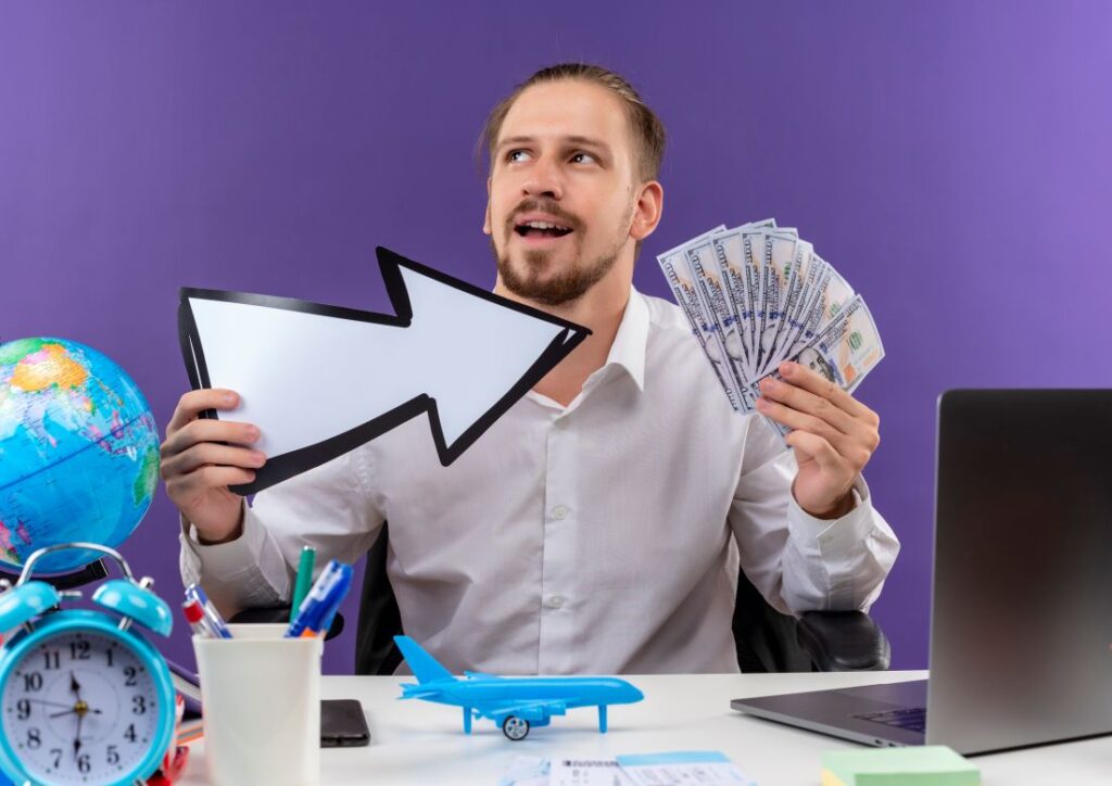 Businessman smiling and holding a white arrow and cash while sitting at a desk, symbolizing revenue growth, business success, and pricing strategies.