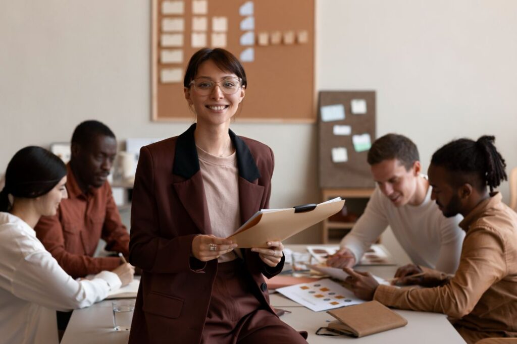 Smiling professional woman standing confidently with a clipboard in a collaborative office environment, with colleagues discussing in the background.